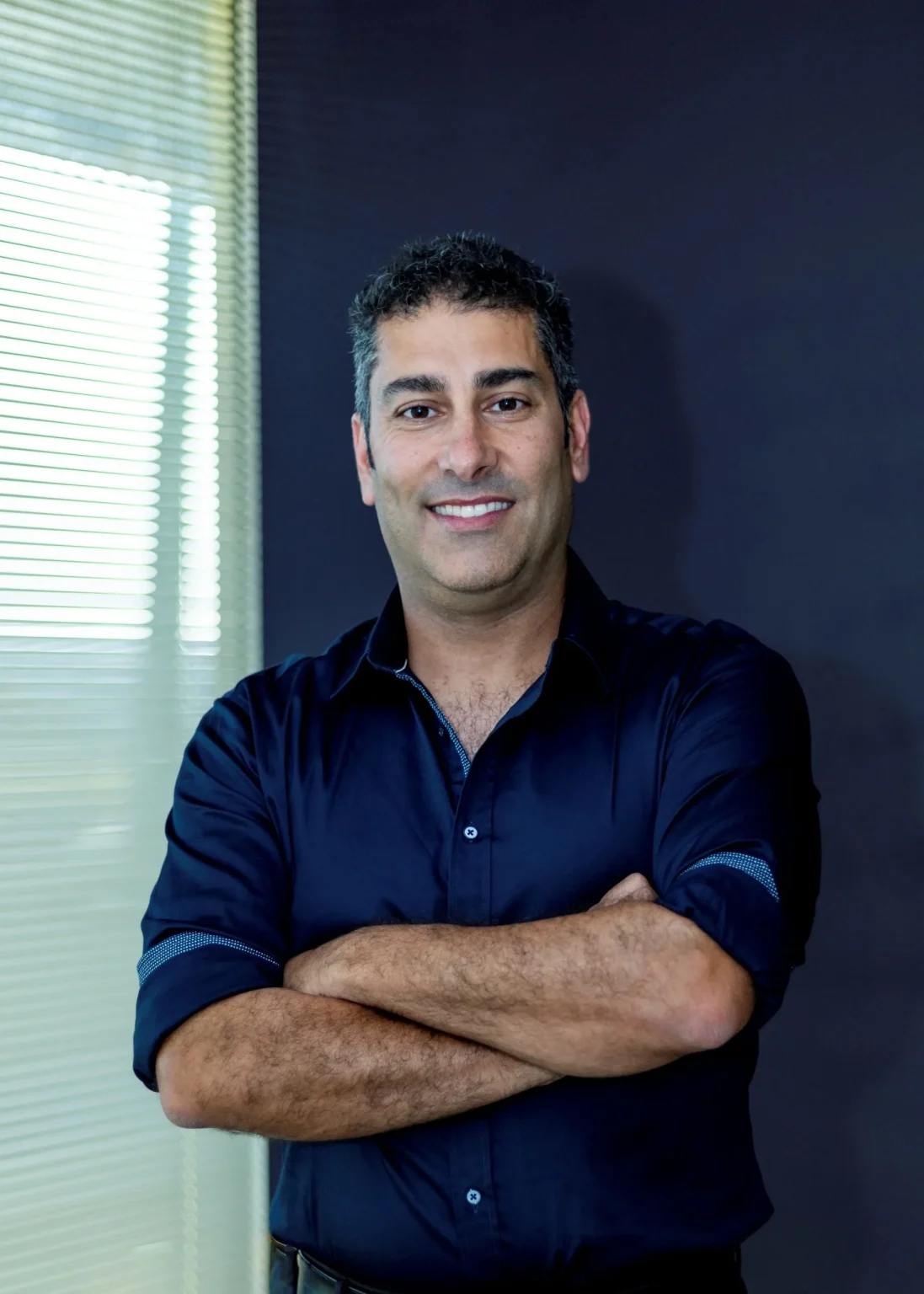 A smiling dentist in Rockingham wearing a dark blue shirt, standing confidently with arms crossed in a professional setting.